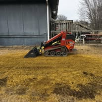 Mini track loader grading and seeding dirt near a barn